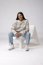 Load image into Gallery viewer, She is sitting laughing wearing a cream colored merino wool chunky knit sweater with green purple and pink jacquard patterns
